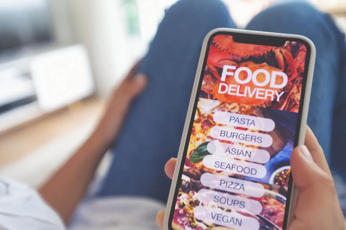 Woman holding a mobile phone with a food delivery menu on it. She is ordering her meal from a menu including pizza, pasta, burgers etc.